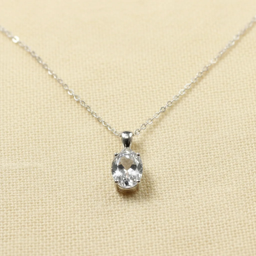 A silver chain necklace with an oval-cut clear gemstone pendant, displayed on a warm cream fabric texture background with text