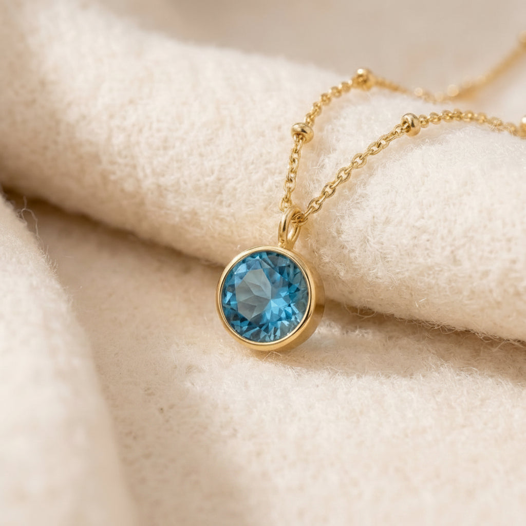 By Sophi Blue Topaz Birthstone Pendant Necklace flat-lay on warm cream wool