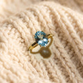 By Sophi Natural Blue Topaz Solitaire Ring in Gold, flat-lay on warm cream wool