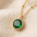 By Sophi Emerald Birthstone Pendant Necklace flat-lay on warm cream wool