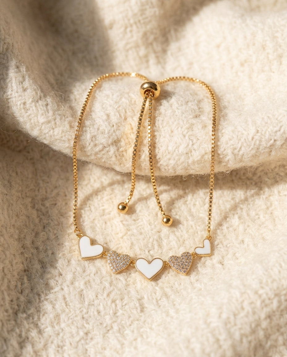 By Sophi White Enamel & Gold Heart Bracelet flat-lay on warm cream wool